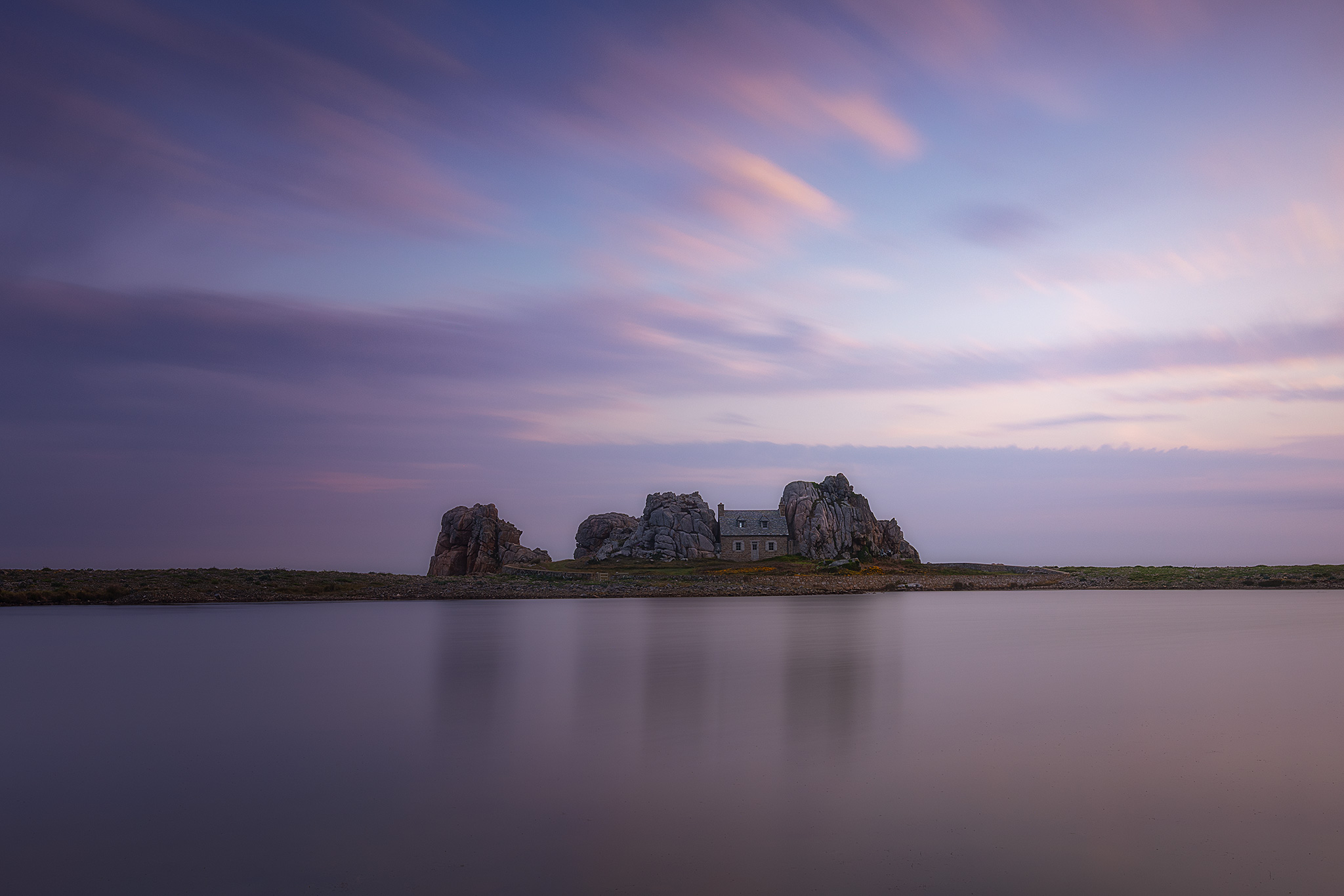 cushy Pose longue devant le Castel Meur en Bretagne, maison en pierre nichée entre des rochers sur un plan d’eau calme à l’heure bleue.