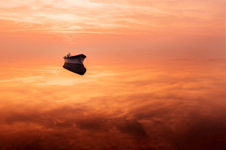 Une barque solitaire repose sur l'eau immobile de l'étang de Canet-en-Roussillon, dans les Pyrénées-Orientales, entièrement embrasée par les teintes orangées du coucher de soleil. Le reflet parfait du ciel dans l'eau brouille la limite entre les deux éléments. Barque solitaire sur une eau immobile, coucher de soleil orangé, étang de Canet-en-Roussillon, Pyrénées-Orientales.
