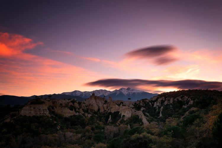Le ciel s'embrase de teintes roses et orangées au-dessus des Orgues d'Ille-sur-Têt, ces cheminées de fée sculptées par l'érosion. En arrière-plan, le Canigó affiche ses sommets enneigés, surmonté de nuages lenticulaires étirés par la pose longue. Coucher de soleil sur les Orgues d'Ille-sur-Têt et le Canigó enneigé, Pyrénées-Orientales.
