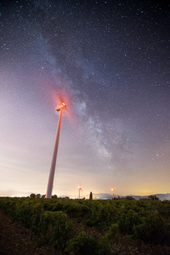 Vent interstellaire. Une éolienne sous la Voie lactée. Éolienne avec balisage lumineux rouge sous la Voie lactée, vignoble en premier plan.